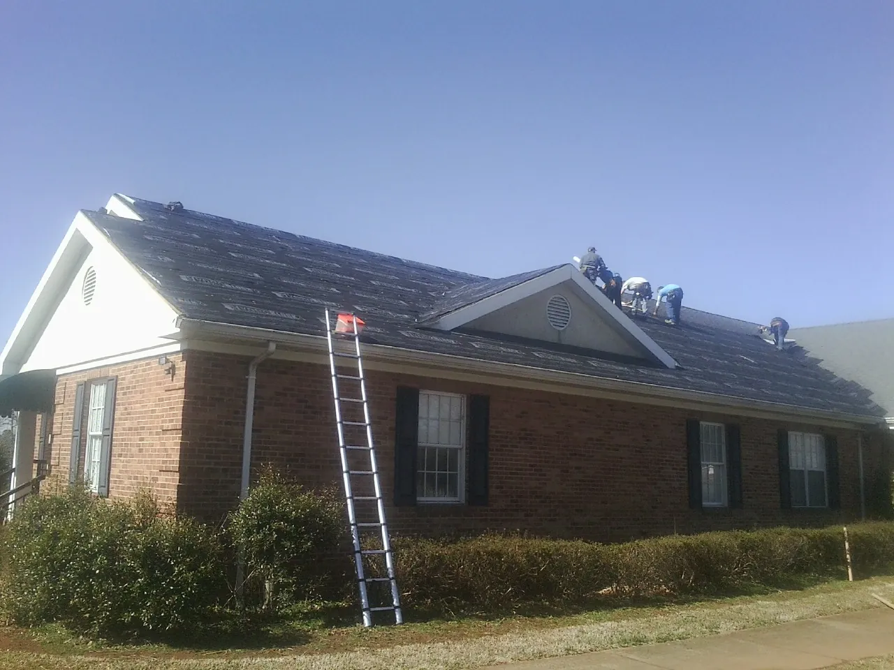 Crew tearing shingles off a brick ranch roof with a ladder set — Emergency Roof Repair work by our crew in Rippey
