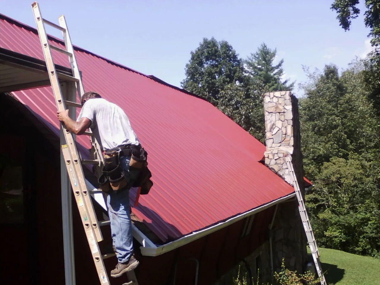 Roofer on a ladder beside a red metal roof and stone chimney — Soffit & Fascia Repair by Roofing Brothers in Rippey